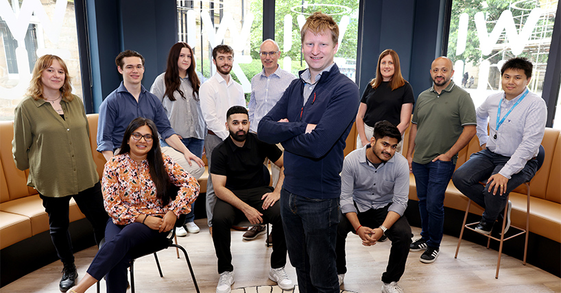 Group from Kinewell Energy sitting in semi circle around Dr Andrew Jenkin who is standing with arms crossed.